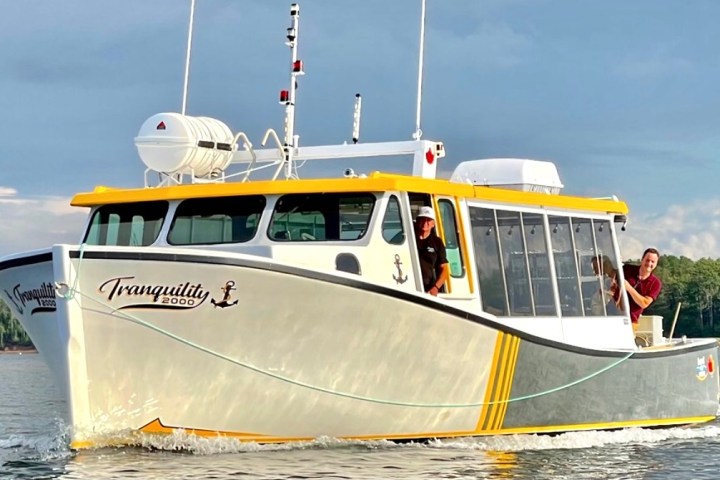 Boat named Tranquility 2000 with a yellow roof on water, people visible, trees in the background.